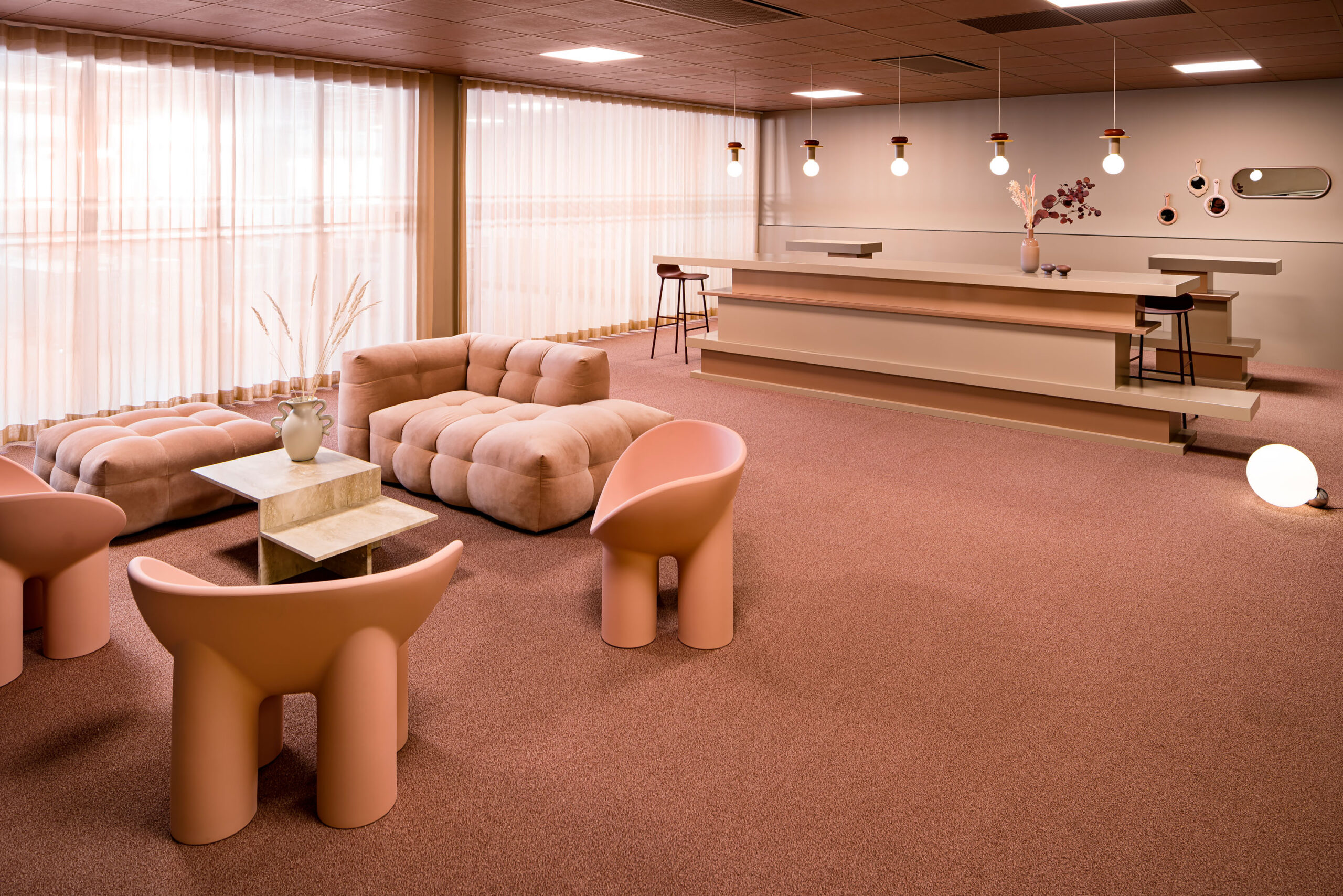 Interior design concept for an office with pink and beige as the main colors. A cozy lounge area with three Roly Poly chairs from Studio Pomone and a Canapé Kendall sofa, a standalone divan from Mogihome in a pink color. Distinct coffee table from Danish brand Ferm Living, made of travertine. Custom-designed bar counter in varying shades of beige and pink.