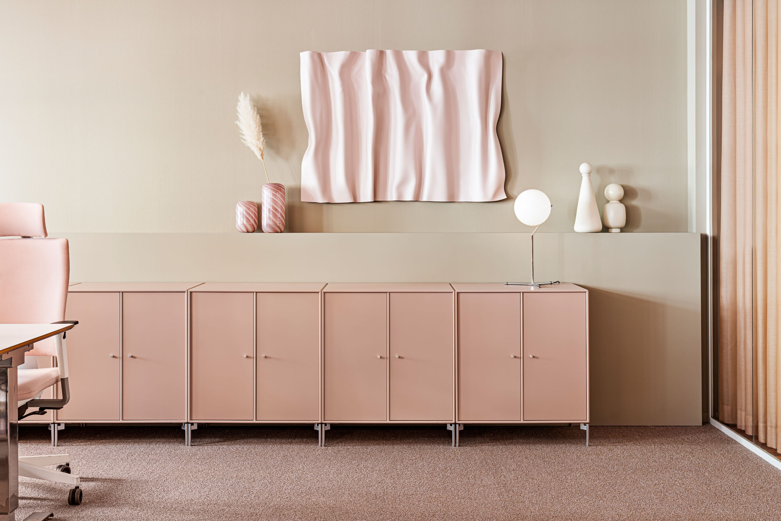Office interior for Saether, with an open plan layout in beige and pink. A view towards a wall where the color scheme is beige, with interior details in different shades of pink. Cabinets, office chairs, vases, a painting, curtains, and a lamp in various shades of pink.